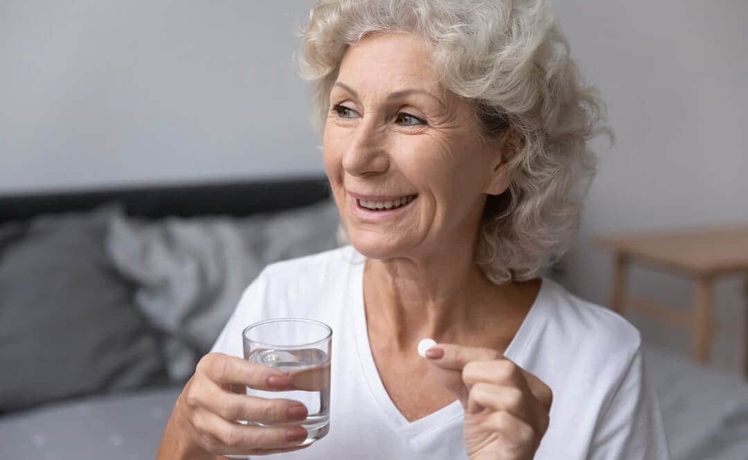 A smiling senior woman with short gray hair holds a pill in one hand and a glass of water in the other