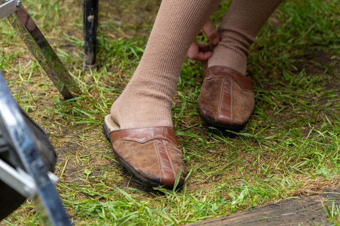 Close-up of an elderly person's feet wearing loose-fitting brown slip-on shoes and tan socks outdoors on grass, with a walker nearby
