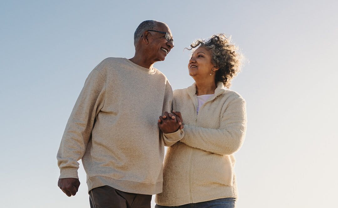 A happy senior couple holds hands and smiles at each other while walking outdoors against a clear blue sky