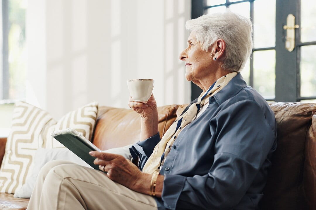 An elegant senior woman with short white hair sits on a leather sofa, holding a cup of tea and a tablet while gazing out a sunlit window